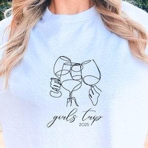 May include: White t-shirt with a black line drawing of three wine glasses and the text "girls trip 2023"