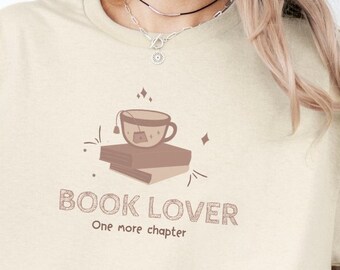 May include: A beige short-sleeve t-shirt featuring a graphic of a teacup resting on two stacked books. The text "BOOK LOVER" is printed in large letters above the smaller text "One more chapter," accompanied by small star and dash accents.