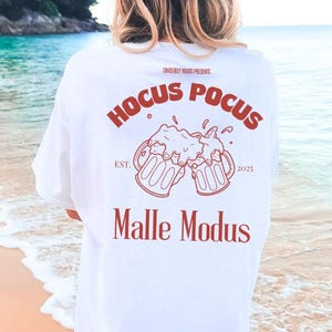 May include: White t-shirt with a red graphic design featuring two beer mugs clinking together. The text "Hocus Pocus" is above the mugs and "Malle Modus" is below. The text "EST. 2023" is between the mugs.