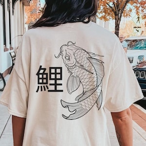 Könnte beinhalten: Ein cremefarbenes T-Shirt mit kurzen Ärmeln und einer schwarzen Strichgrafik eines Koi-Karpfens auf der Rückseite. Links neben dem Fisch ist das japanische Kanji-Zeichen für Koi in Schwarz aufgedruckt. Das Shirt hat einen Rundhalsausschnitt und eine lockere Passform.