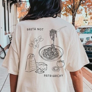 May include: An off-white short-sleeve t-shirt featuring a black line art graphic on the back. The design includes illustrations of pasta, a grater, cheese, and a tomato, with the text "PASTA NOT PATRIARCHY" printed in a simple sans-serif font.