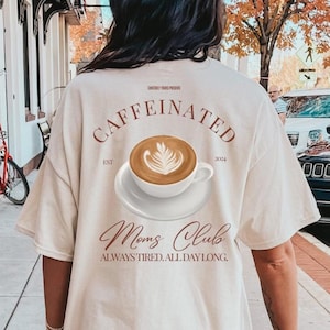 Op de afbeelding: Een crèmekleurig t-shirt met een afbeelding van een koffiekopje met latte art op de achterkant. De tekst luidt CAFFEINATED, EST 2024, Moms Club en ALWAYS TIRED. ALL DAY LONG. Het ontwerp is in bruine inkt op de achterkant van het t-shirt gedrukt.