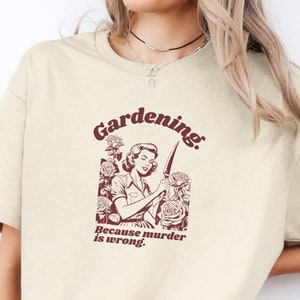 Könnte beinhalten: Ein beiges T-Shirt mit kurzen Ärmeln und einem dunkelroten Grafikdruck einer Person, die ein Messer zwischen Rosen hält. Der Text über der Abbildung lautet Gardening. und der Text darunter lautet Because murder is wrong.