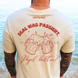 May include: A cream colored t-shirt with a red graphic on the back. The graphic features two beer mugs clinking together with the text "Egal Was Passiert" above and "Pegel halten!" below. The text "EST. 2023" is also included in the graphic.