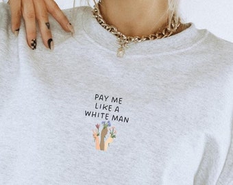 Puede incluir: Una sudadera gris claro de cuello redondo con el texto negro PAY ME LIKE A WHITE MAN centrado en el pecho. Debajo del texto hay una pequeña ilustración gráfica de dos manos de diferentes tonos de piel sosteniendo flores coloridas.