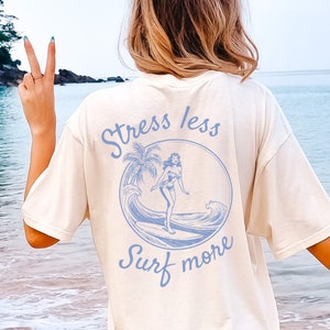Könnte beinhalten: Ein weißes T-Shirt mit einer blauen Grafik einer Frau beim Surfen mit dem Text "Stress less Surf more" auf der Rückseite.