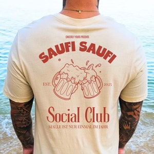 May include: A beige t-shirt with a red graphic design on the back. The graphic features two beer mugs clinking together with the text "Saufi Saufi" above and "Social Club" below. The text "Est. 2025" and "Malle ist nur einmal im Jahr" are also included in the design.