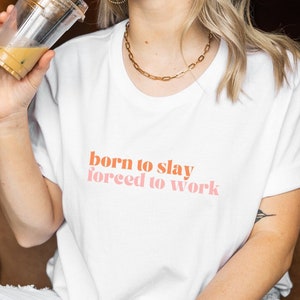 Könnte beinhalten: Weißes T-Shirt mit dem Text "born to slay forced to work" in orangefarbener und rosafarbener Schrift.