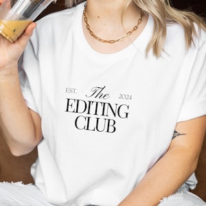 Könnte beinhalten: Ein weißes T-Shirt mit schwarzem Text, der "EST. 2024 The Editing Club" lautet. Die Person, die das Shirt trägt, lächelt und hält eine Tasse Eiskaffee.