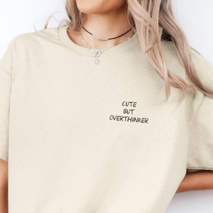 Könnte beinhalten: Ein beiges T-Shirt mit dem Aufdruck "Cute but Overthinker" auf der Vorderseite.