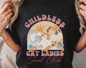 May include: A black t-shirt featuring a graphic print of three cats in front of a sunset and palm trees. The text reads CHILDLESS CAT LADIES SOCIAL CLUB in pink and orange lettering, accented with colorful tropical flowers at the base.