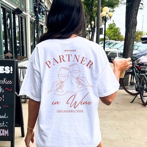 May include: White t-shirt with a red graphic of two wine glasses clinking. The text "PARTNER in Wine" is written below the glasses. The text "LISA SANDRA ANNE" is written below the text "in Wine".