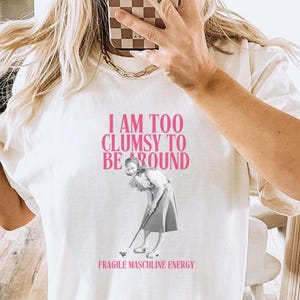 Puede incluir: Camiseta blanca con un gráfico rosa que dice "Soy demasiado torpe para estar cerca" y una imagen en blanco y negro de una mujer jugando al golf. El texto "Fragile Masculine Energy" está impreso debajo de la imagen.