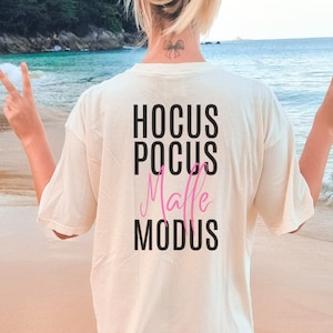 Può includere: Una maglietta bianca con la scritta "HOCUS POCUS Malle MODUS" stampata sul retro. La scritta "Malle" è in rosa.