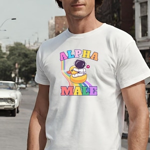 Könnte beinhalten: Weißes T-Shirt mit einem Aufdruck eines Astronauten, der auf einer regenbogenfarbenen Gummi-Ente reitet. Der Text "ALPHA MALE" ist unter dem Aufdruck gedruckt.