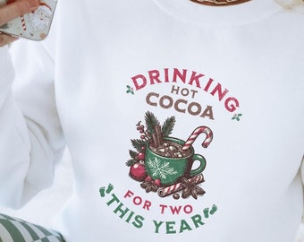 May include: White sweatshirt with a Christmas-themed graphic that says "Drinking Hot Cocoa For Two This Year". The graphic features a green mug with hot cocoa, marshmallows, a candy cane, and other festive decorations.