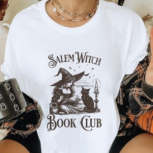 May include: White t-shirt with a black and white illustration of a witch reading a book with a black cat. The text "Salem Witch Book Club" is printed on the shirt.