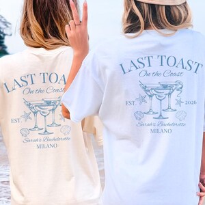 Könnte beinhalten: Zwei kurzärmelige T-Shirts, eines in Creme und eines in Weiß, mit einer blauen Grafik auf der Rückseite. Das Design enthält den Text LAST TOAST On the Coast, EST. 2026, Sarah's Bachelorette MILANO sowie Abbildungen von Martinigläsern, Muscheln und Seesternen.