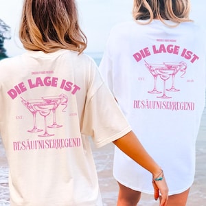 May include: A cream-colored t-shirt and a white t-shirt, each featuring a pink graphic on the back. The design includes illustrations of three cocktail glasses and the text "DIE LAGE IST BESÄUFNISERREGEND" along with "EST. 2026" and "UNSERE VIBES PRESENTS".