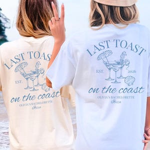 Puede incluir: Dos camisetas de manga corta, una color crema y otra blanca, con un gráfico azul en la espalda. El diseño incluye dos copas de cóctel, una sombrilla de playa, un caballito de mar y una concha, con el texto LAST TOAST on the coast, EST. 2026, OLIVIA'S BACHELORETTE e Ibiza.