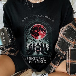 May include: A black t-shirt featuring a graphic print of four witches gathered around a cauldron under a large red moon. The text above reads WITH A LITTLE EXTRA MAGIC and the text below reads GIRLS WILL BE GIRLS. The shirt is worn with a plaid shirt.