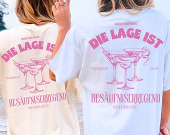 May include: Two short-sleeve t-shirts, one cream and one white, featuring pink graphic prints on the back. The design includes cocktail glasses, the text "DIE LAGE IST BESÄUFNISERREGEND", "Budapest", "2026", and "SIE HEIRATET!" or "ICH HEIRATE!".