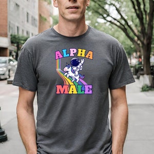 Könnte beinhalten: Ein graues T-Shirt mit dem Text "ALPHA MALE" in Regenbogenfarben. Ein Astronaut reitet auf einem Einhorn auf einem Regenbogen.