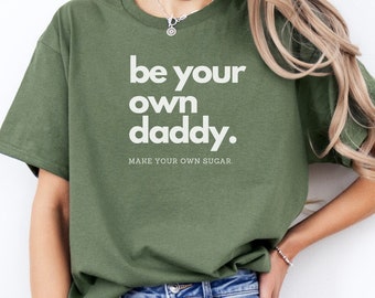 May include: An olive green short-sleeved t-shirt featuring the white text "be your own daddy." printed in a bold sans-serif font, with the smaller white text "MAKE YOUR OWN SUGAR." positioned directly underneath the main phrase.