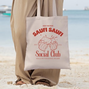 May include: A white canvas tote bag with red text and a graphic of two beer mugs clinking. The text reads "Saufi Saufi Social Club Est. 2025 Malle ist nur einmal im Jahr."