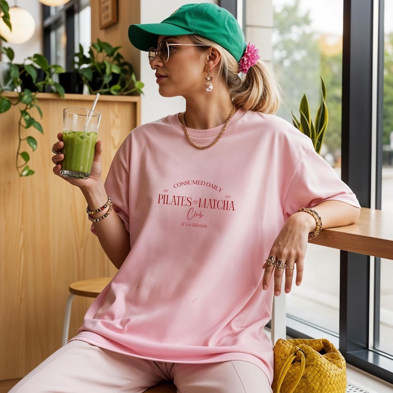May include: A light pink t-shirt with the text "CONSUMED DAILY PILATES & MATCHA Club it's a lifestyle" in red. The person is wearing a green baseball cap, gold jewelry, and holding a glass of green drink.