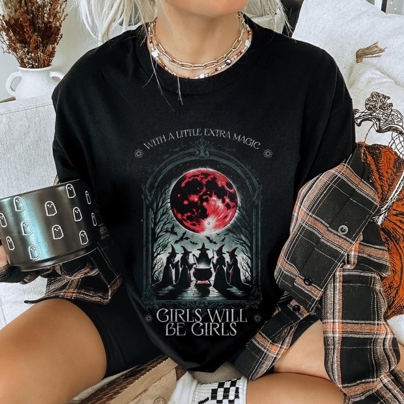 May include: A black t-shirt featuring a graphic print of four witches gathered around a cauldron under a large red moon. The text above reads WITH A LITTLE EXTRA MAGIC and the text below reads GIRLS WILL BE GIRLS. The shirt is worn with a plaid shirt.