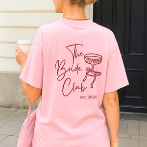 May include: A light pink t-shirt with the text "The Bride Club" and a champagne glass illustration in burgundy. The shirt also includes "est. 2026". The person is holding a white coffee cup and a pink bag.
