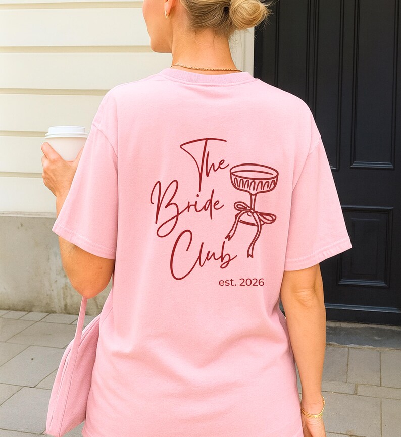 May include: A light pink t-shirt with the text "The Bride Club" and a champagne glass illustration in burgundy. The shirt also includes "est. 2026". The person is holding a white coffee cup and a pink bag.