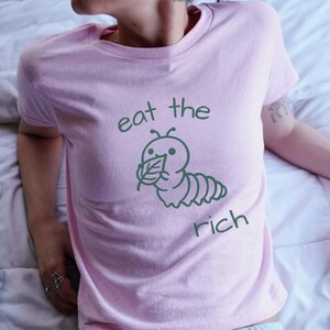 May include: A light pink short-sleeved t-shirt featuring a dark green graphic of a caterpillar eating a leaf. The text "eat the" is printed above the caterpillar, and the word "rich" is printed below it in a matching dark green font.