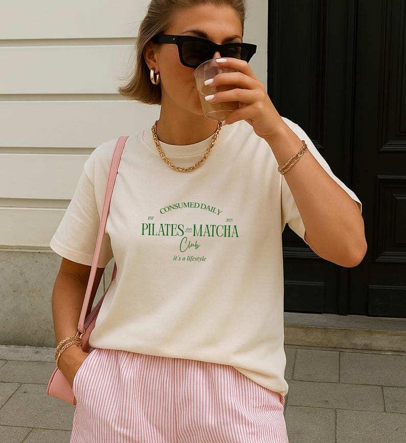 May include: Cream-colored t-shirt with the text "CONSUMED DAILY PILATES and MATCHA Club" in green. The shirt is paired with pink and white striped shorts and a pink shoulder bag. The person is wearing gold jewelry and black sunglasses.