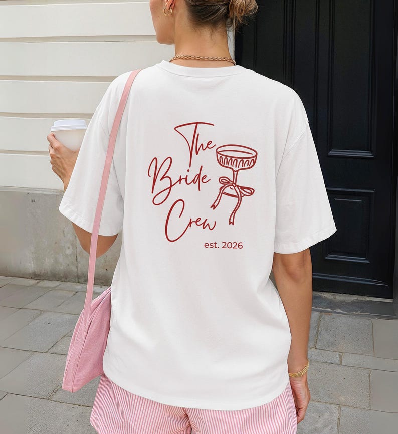 May include: A white t-shirt with the text "The Bride Crew" in a burgundy script font, along with a matching graphic of a cocktail glass and the year "est. 2026". The shirt is part of a bridal party theme.