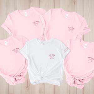 Pode incluir: T-shirts rosa e brancas com o texto "Team Bride" e "The Bride" em fonte cursiva. As camisas apresentam um gráfico de taça de champanhe. Algumas camisas têm o texto "Sophia's Bach Party" na manga.