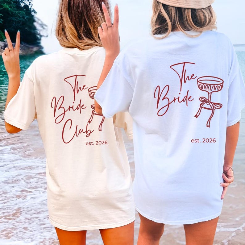 May include: Two t-shirts, one cream and one white, with the text "The Bride Club" and "est. 2026" in burgundy script. The white shirt features a graphic of a champagne glass. The shirts are worn on a beach.