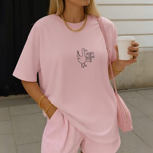 May include: A light pink t-shirt with a cartoon duck holding a sign that reads "BURN THE PATRIARCHY." The shirt is paired with pink and white striped shorts. A gold chain necklace and bracelet are visible.