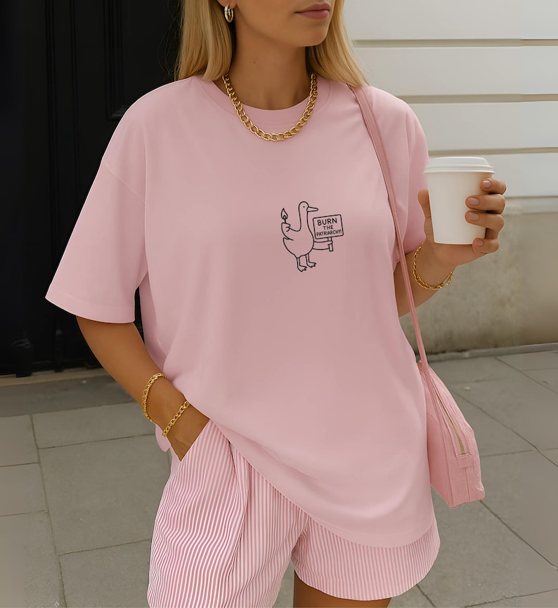 May include: A light pink t-shirt with a cartoon duck holding a sign that reads "BURN THE PATRIARCHY." The shirt is paired with pink and white striped shorts. A gold chain necklace and bracelet are visible.