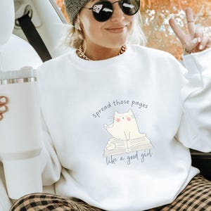 May include: A white crewneck sweatshirt featuring a graphic of a cream-colored cat sitting on an open book. The text above the cat reads "spread those pages" and the text below reads "like a good girl" in a simple, dark blue sans-serif font.