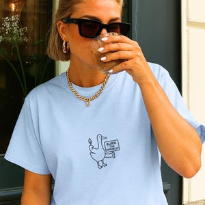 May include: Light blue t-shirt featuring a black and white graphic of a duck holding a candle and a sign that reads "BURN THE PATRIARCHY!". The shirt is made of a soft material.