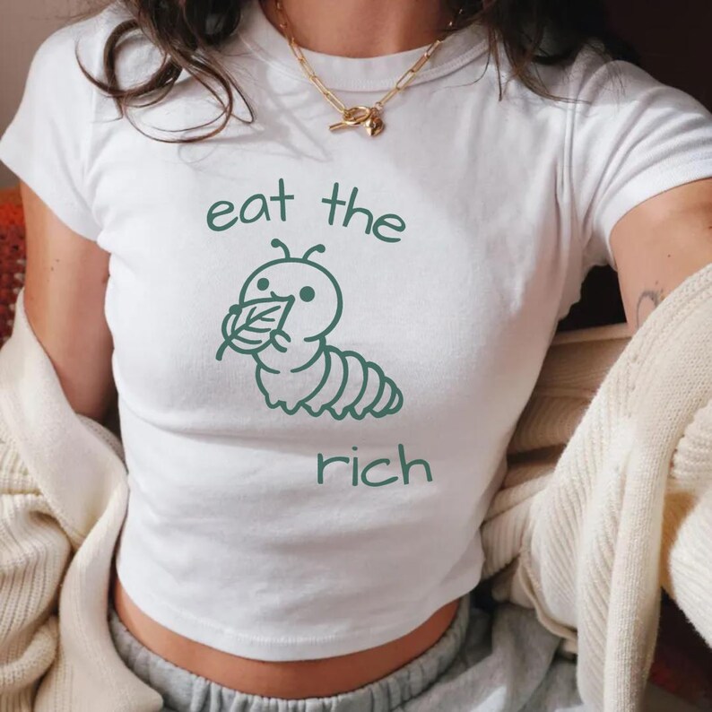 May include: A white short-sleeved t-shirt featuring a dark green graphic of a caterpillar eating a leaf. The text "eat the" is printed above the caterpillar and the word "rich" is printed below it in a matching dark green font.