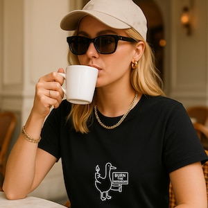 May include: A black t-shirt with a white graphic of a duck holding a lit match and a sign that reads "BURN THE PATRIARCHY!". The person is wearing a beige baseball cap, black sunglasses, and gold jewelry, and is holding a white mug.