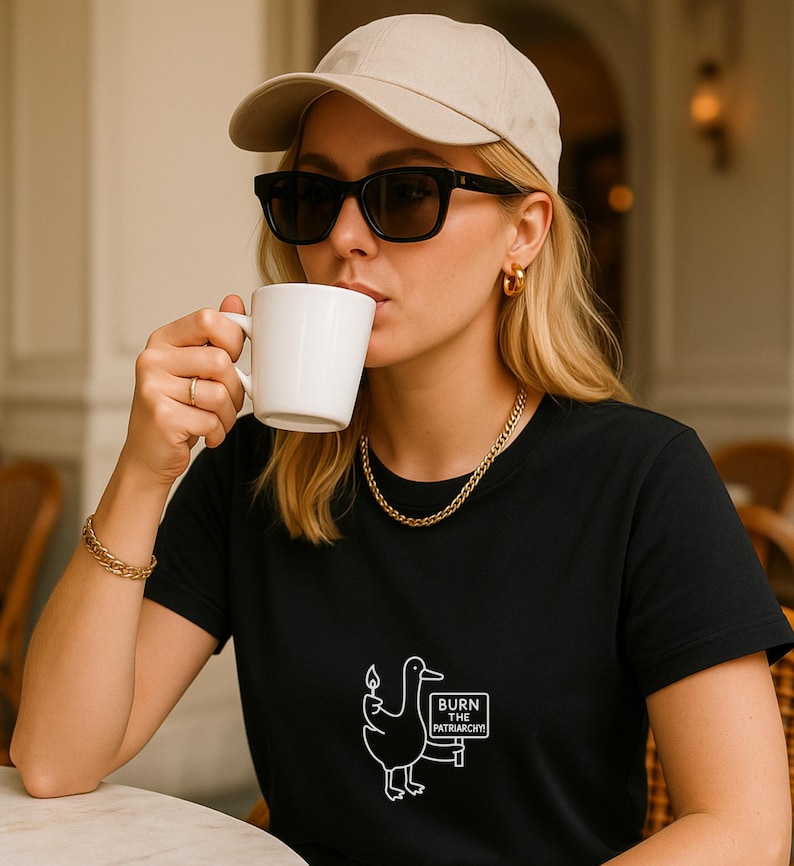 May include: A black t-shirt with a white graphic of a duck holding a lit match and a sign that reads "BURN THE PATRIARCHY!". The person is wearing a beige baseball cap, black sunglasses, and gold jewelry, and is holding a white mug.