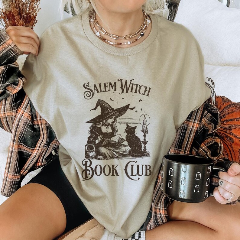 May include: A light beige t-shirt with a vintage illustration of a witch reading a book with a black cat beside her. The text "Salem Witch Book Club" is printed on the shirt.