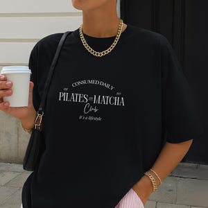 May include: Black oversized t-shirt with the text "CONSUMED DAILY PILATES & MATCHA Club" in white. The shirt has a crew neck and short sleeves. A gold chain necklace and bracelet are also visible.