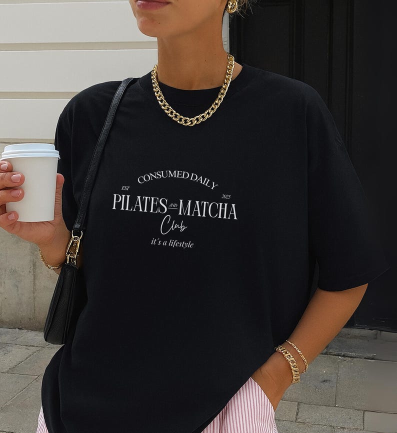 May include: Black oversized t-shirt with the text "CONSUMED DAILY PILATES & MATCHA Club" in white. The shirt has a crew neck and short sleeves. A gold chain necklace and bracelet are also visible.