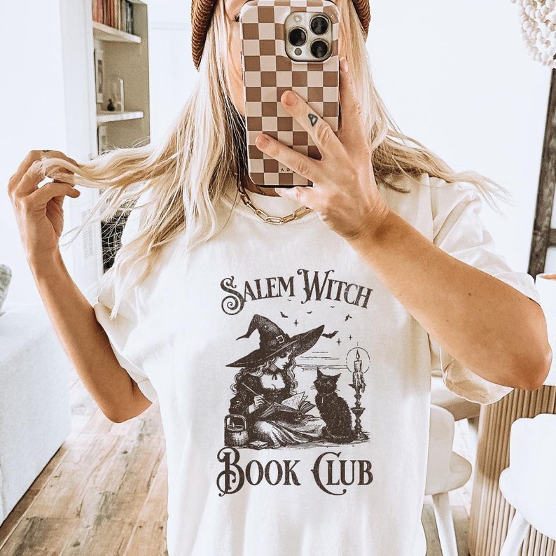 May include: A white t-shirt with a black and white illustration of a witch reading a book with a black cat sitting next to her. The text "Salem Witch Book Club" is printed on the shirt.