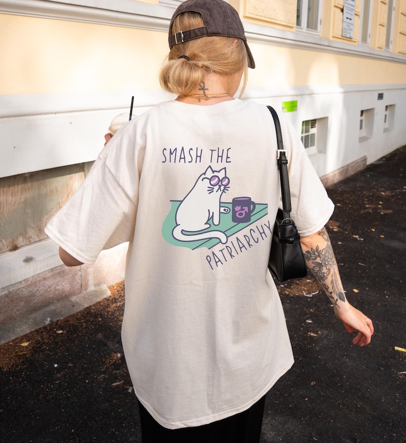 May include: Cream-colored t-shirt with the text "SMASH THE PATRIARCHY" and a graphic of a white cat wearing glasses, sitting next to a mug. The cat is on a green surface. The shirt is a casual, oversized fit.